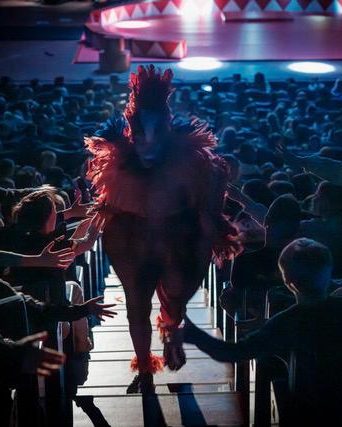 A performer in a red feathered bird costume walks down the aisle through the children in the audience.